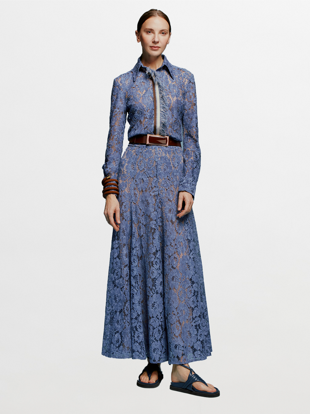 A woman stands against a blue background wearing the Lavanda Skirt Blue paired with a long-sleeved, blue lace top, accessorized with a brown belt, beaded necklace, and brown bangles. She has her hair tied back and is wearing black sandals. This stylish look will be available starting August 19th, 2024.