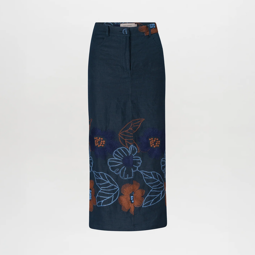 Against a plain background, a woman stands wearing a brown checkered jacket, the Layan Skirt Navy Ochre Flower Embroidery, a brown belt, and gray heeled boots. With her hair tied back neatly, she exudes grace as if ready to board an elegant ship setting sail on August 19th.