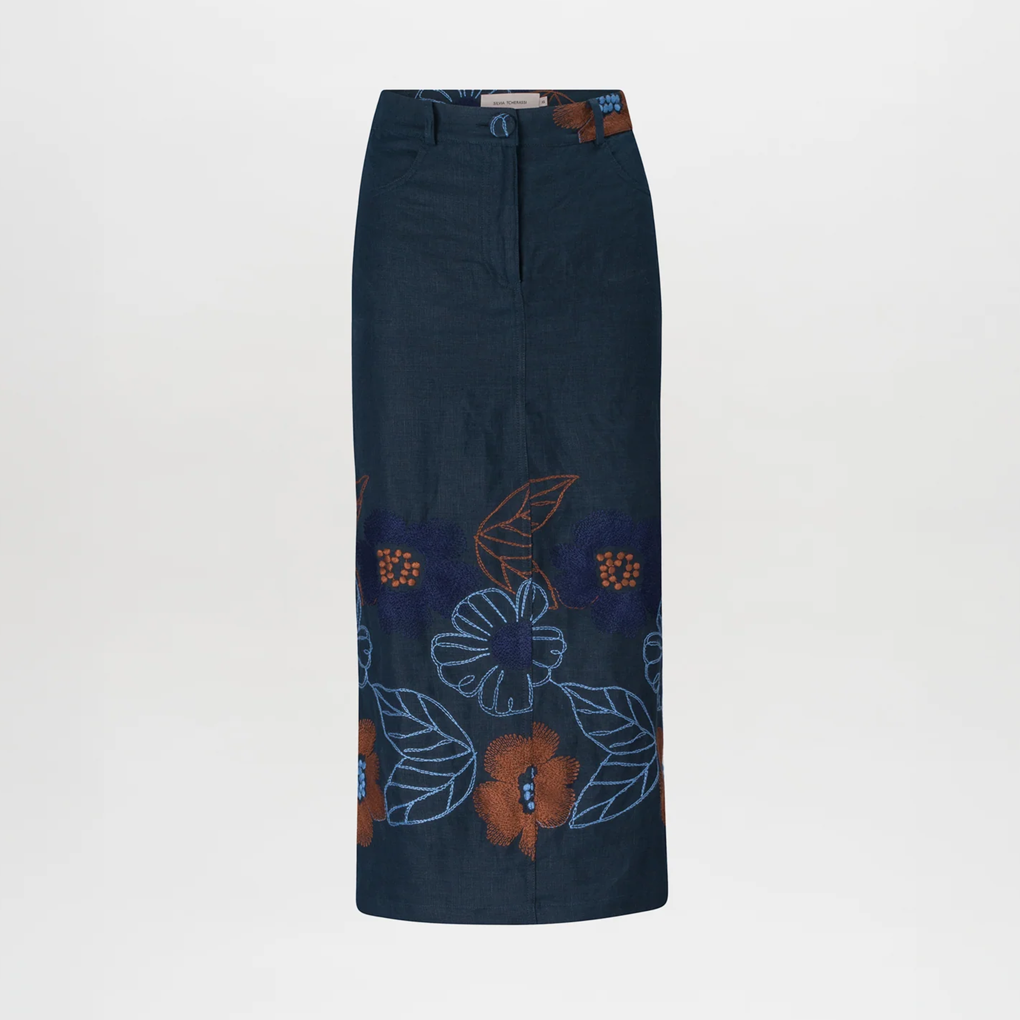 Against a plain background, a woman stands wearing a brown checkered jacket, the Layan Skirt Navy Ochre Flower Embroidery, a brown belt, and gray heeled boots. With her hair tied back neatly, she exudes grace as if ready to board an elegant ship setting sail on August 19th.