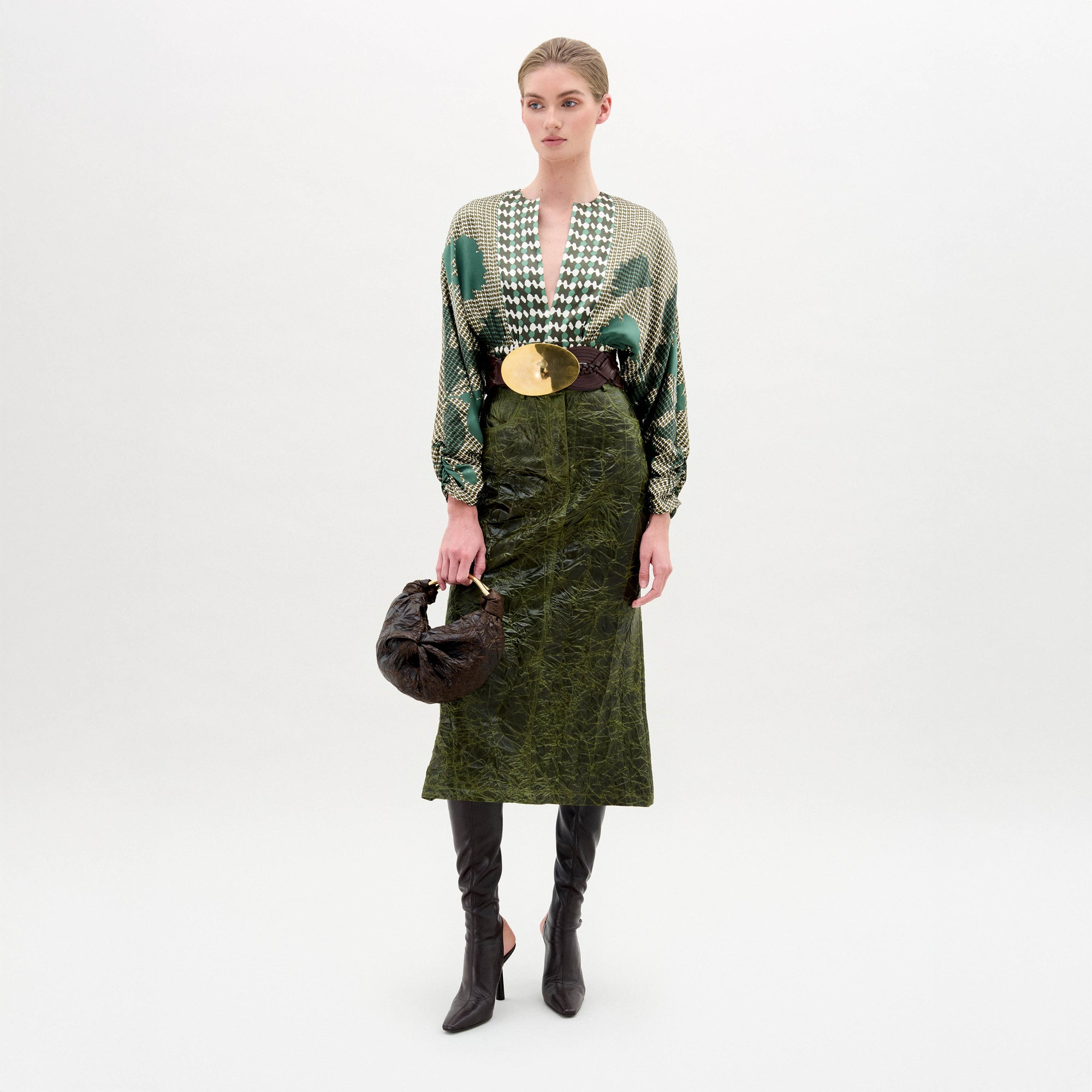 A woman stands against a plain background wearing a patterned green blouse, the Layan Skirt, black boots, and holding a round black bag.