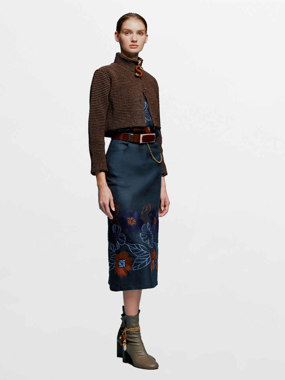 Against a plain background, a woman stands wearing a brown checkered jacket, the Layan Skirt Navy Ochre Flower Embroidery, a brown belt, and gray heeled boots. With her hair tied back neatly, she exudes grace as if ready to board an elegant ship setting sail on August 19th.