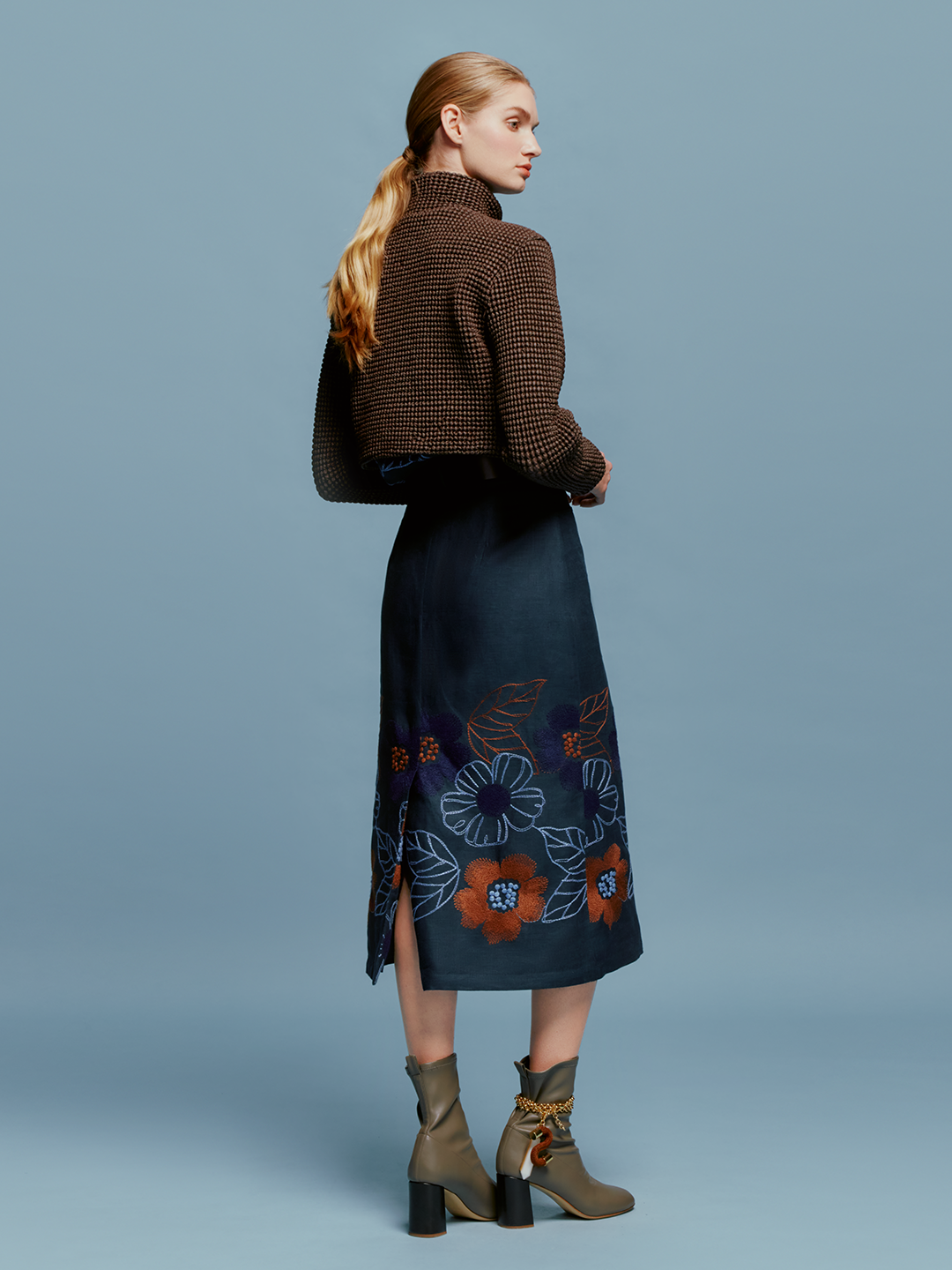 Against a plain background, a woman stands wearing a brown checkered jacket, the Layan Skirt Navy Ochre Flower Embroidery, a brown belt, and gray heeled boots. With her hair tied back neatly, she exudes grace as if ready to board an elegant ship setting sail on August 19th.