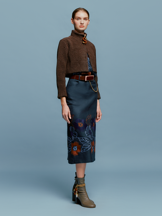 Against a plain background, a woman stands wearing a brown checkered jacket, the Layan Skirt Navy Ochre Flower Embroidery, a brown belt, and gray heeled boots. With her hair tied back neatly, she exudes grace as if ready to board an elegant ship setting sail on August 19th.