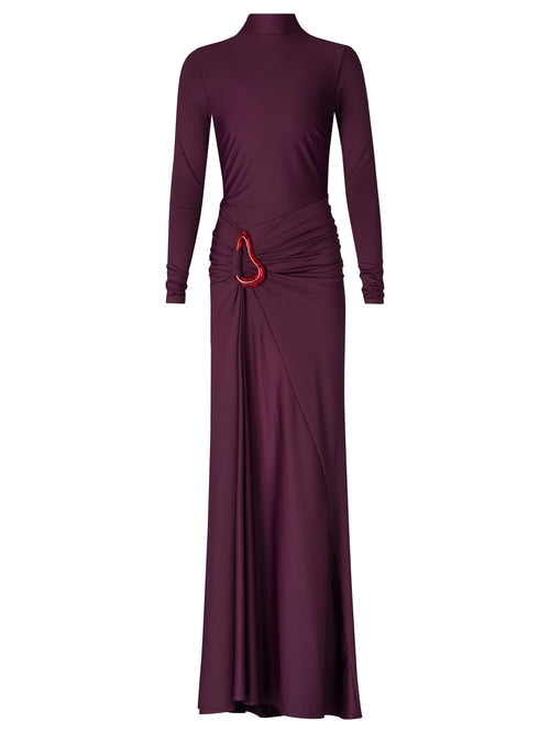 A person in a fitted, long-sleeve Lena Dress Burgundy with a high neckline stands against a plain blue background. The gown, perfect for an August 19th, 2024 gala, features elegant draping and a red embellishment near the waist.