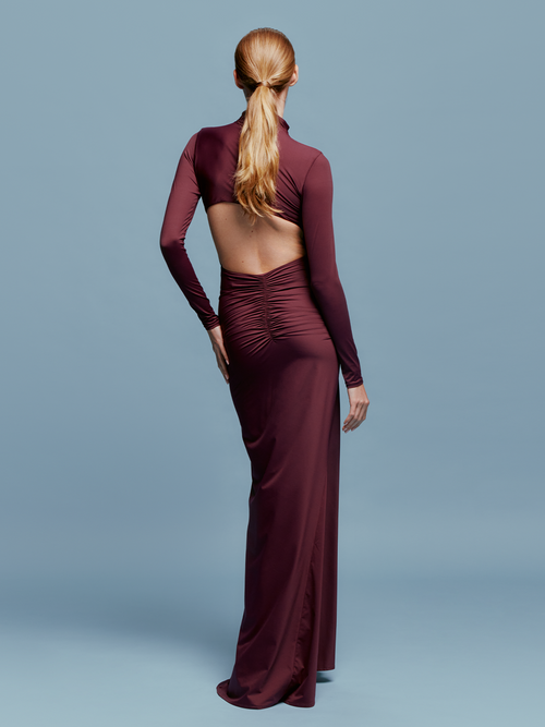 A person in a fitted, long-sleeve Lena Dress Burgundy with a high neckline stands against a plain blue background. The gown, perfect for an August 19th, 2024 gala, features elegant draping and a red embellishment near the waist.