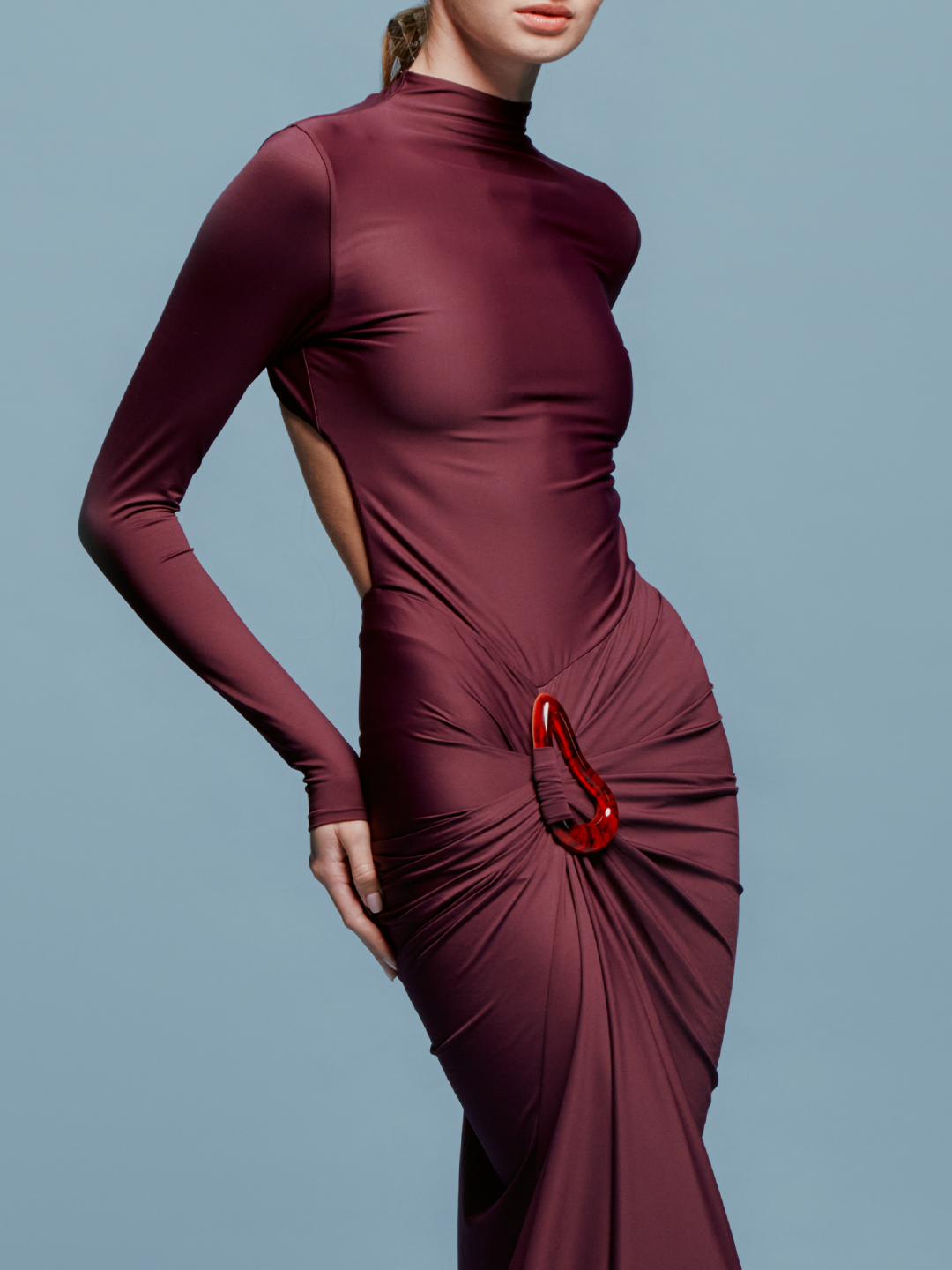 A person in a fitted, long-sleeve Lena Dress Burgundy with a high neckline stands against a plain blue background. The gown, perfect for an August 19th, 2024 gala, features elegant draping and a red embellishment near the waist.