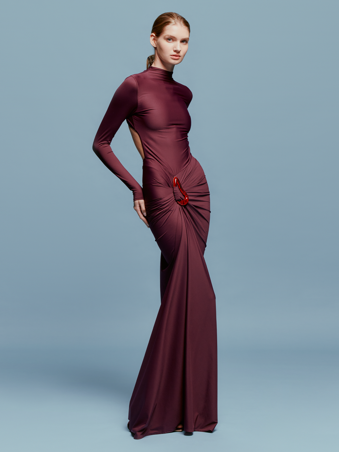 A person in a fitted, long-sleeve Lena Dress Burgundy with a high neckline stands against a plain blue background. The gown, perfect for an August 19th, 2024 gala, features elegant draping and a red embellishment near the waist.