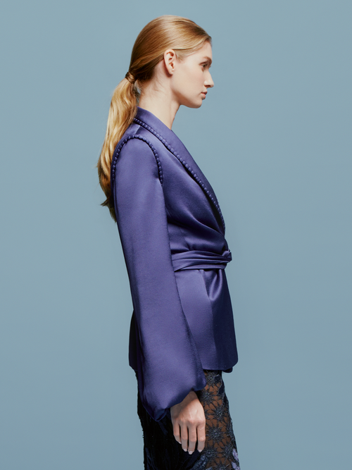 A person in a chic Leo Jacket Navy and a patterned skirt stands against a light blue background, embodying the stylish essence of September 2nd. They are wearing a long necklace with a white pendant.