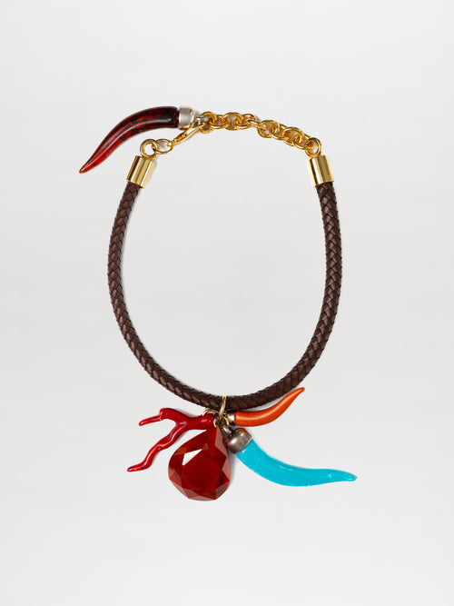 The Leslie Choker is a braided leather piece with metallic gold clasps, featuring a red faceted pendant, a red antler-shaped piece, a red curved horn, and multicolor charms such as a blue curved horn.