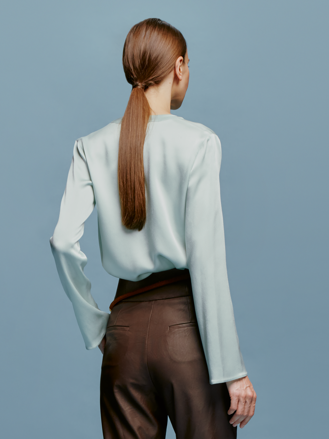 A woman stands against a plain blue background, wearing the Lilly Blouse in Turquoise and brown high-waisted pants, with a relaxed and composed expression, as if ready for September 2nd, 2024.