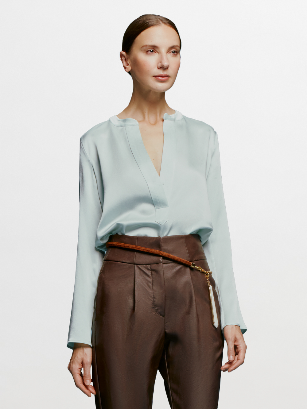 A woman stands against a plain blue background, wearing the Lilly Blouse in Turquoise and brown high-waisted pants, with a relaxed and composed expression, as if ready for September 2nd, 2024.