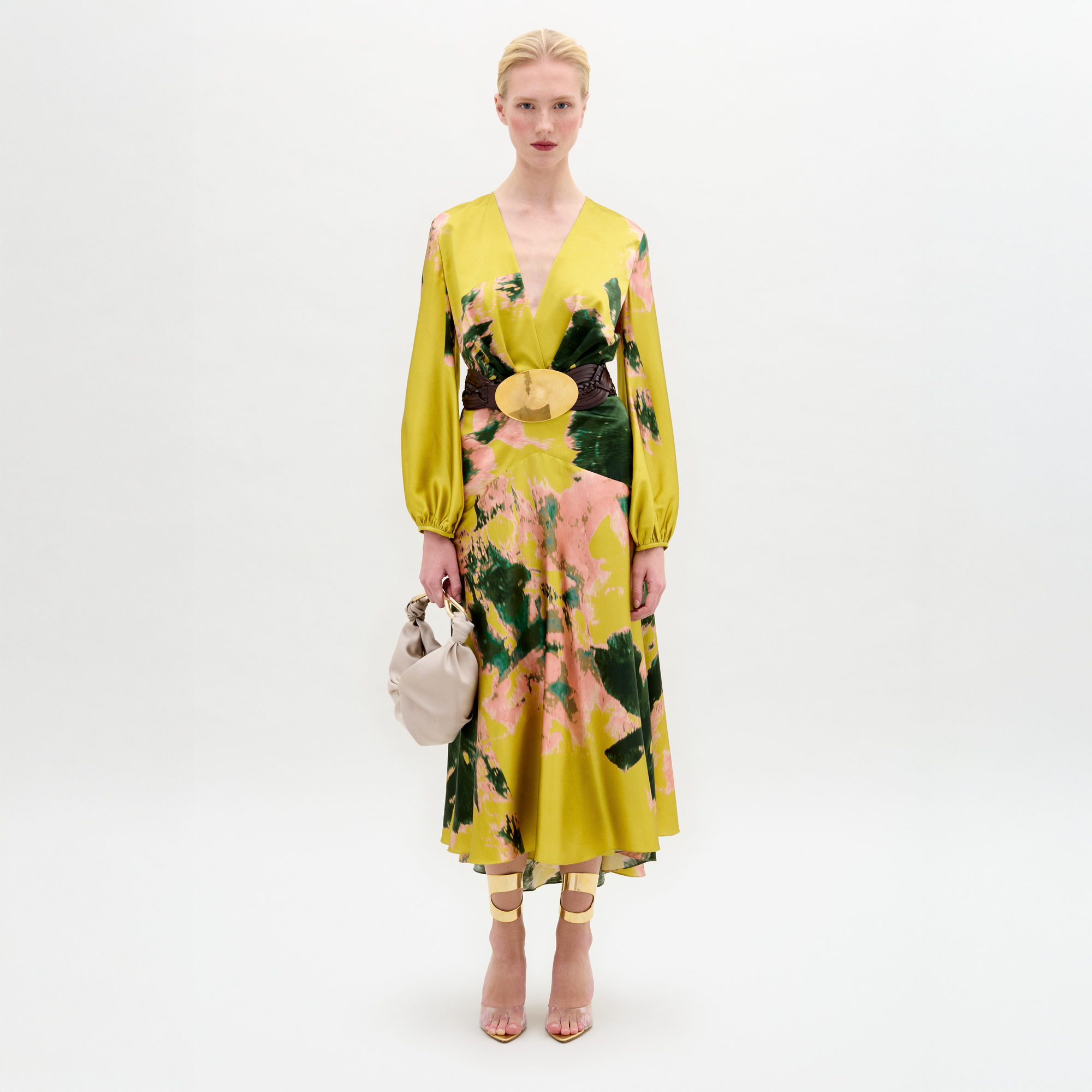 A person in a Lima Dress with a floral yellow and green pattern, a wide belt, holding a beige handbag, and wearing strappy high heels.