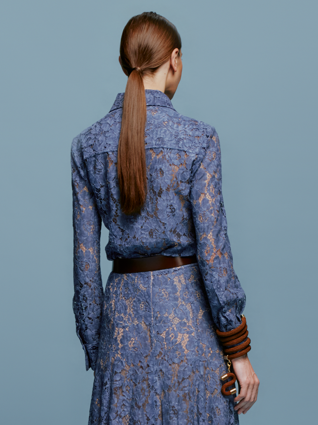 A woman stands against a plain blue background, wearing the Liv Blouse in Blue. She pairs it with a stylish brown bracelet, perfectly capturing the essence of September 2nd, 2024.