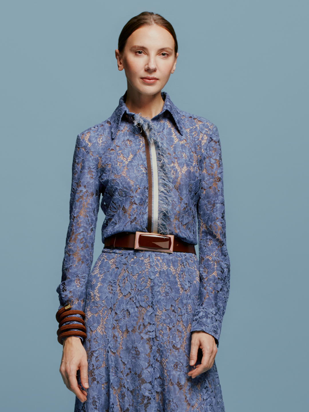 A woman stands against a plain blue background, wearing the Liv Blouse in Blue. She pairs it with a stylish brown bracelet, perfectly capturing the essence of September 2nd, 2024.