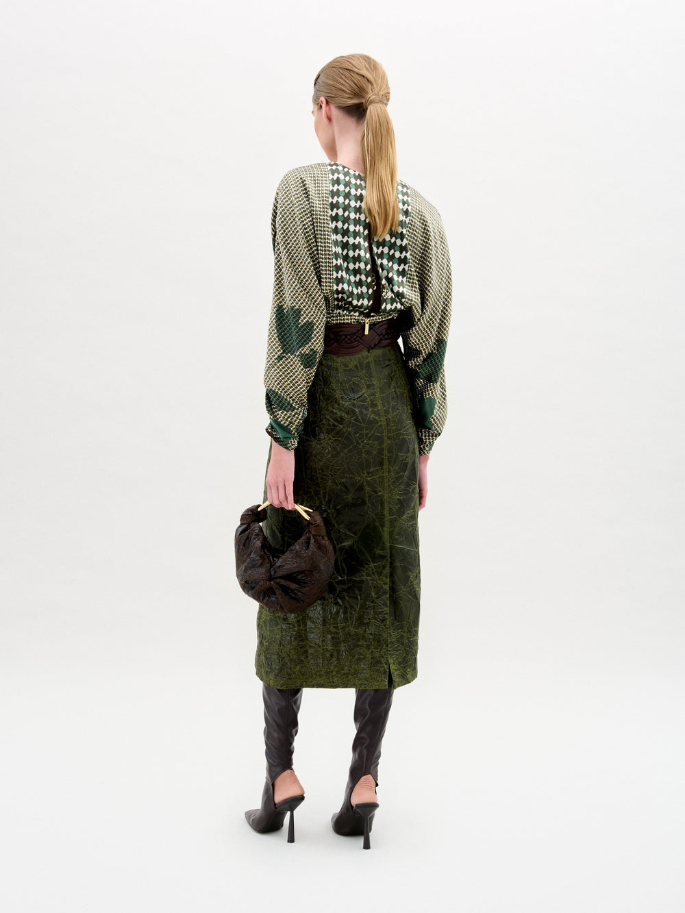 A woman stands against a plain background wearing a patterned green blouse, the Layan Skirt, black boots, and holding a round black bag.