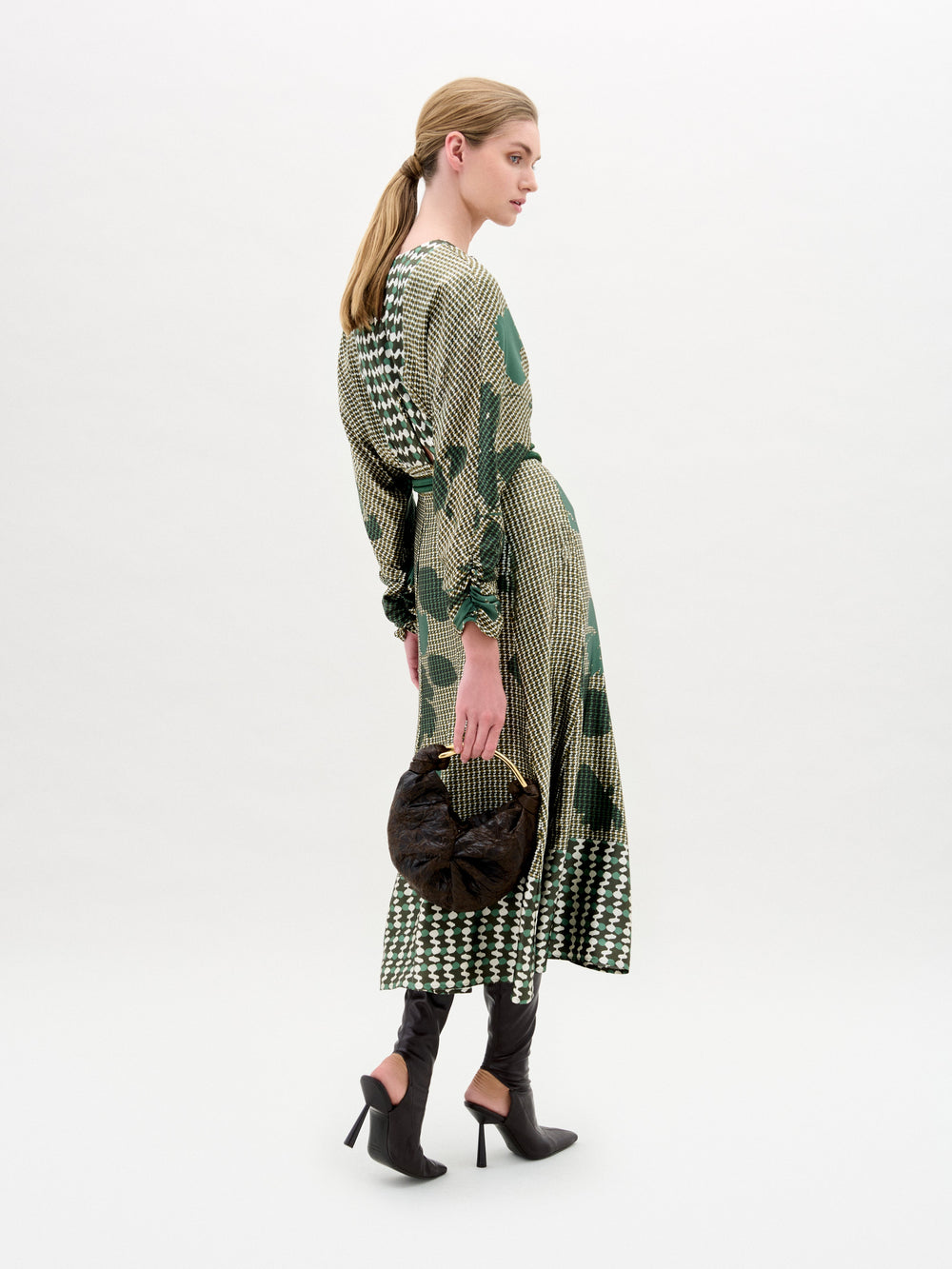 A woman stands against a white background wearing the Lizzy Blouse, a green silk patterned top, paired with a dark green textured skirt, wide belt with large gold buckle, and holding a brown bag.