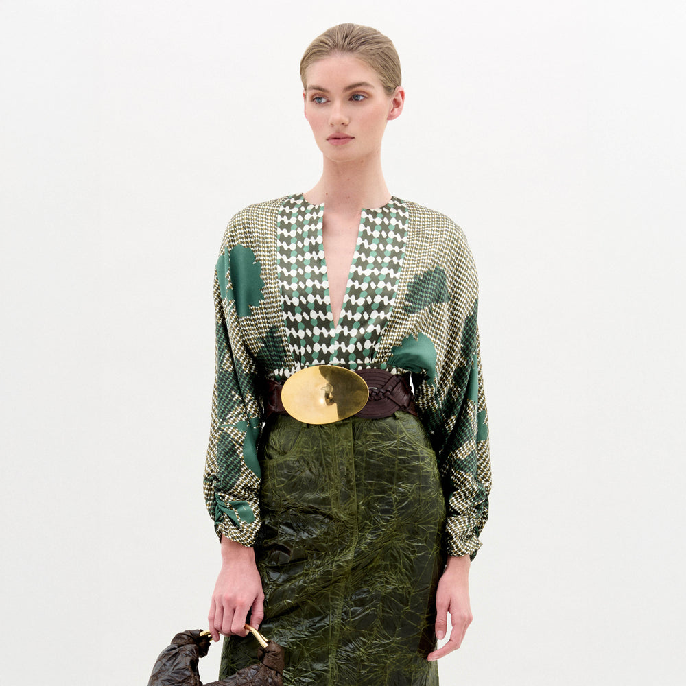 A woman stands against a white background wearing the Lizzy Blouse, a green silk patterned top, paired with a dark green textured skirt, wide belt with large gold buckle, and holding a brown bag.