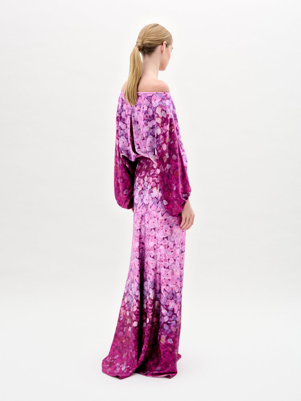 Woman models the Lorella Dress—an off-the-shoulder maxi with purple and pink florals, crafted from luxurious Italian fabric and finished with a tied belt—against a plain white background.