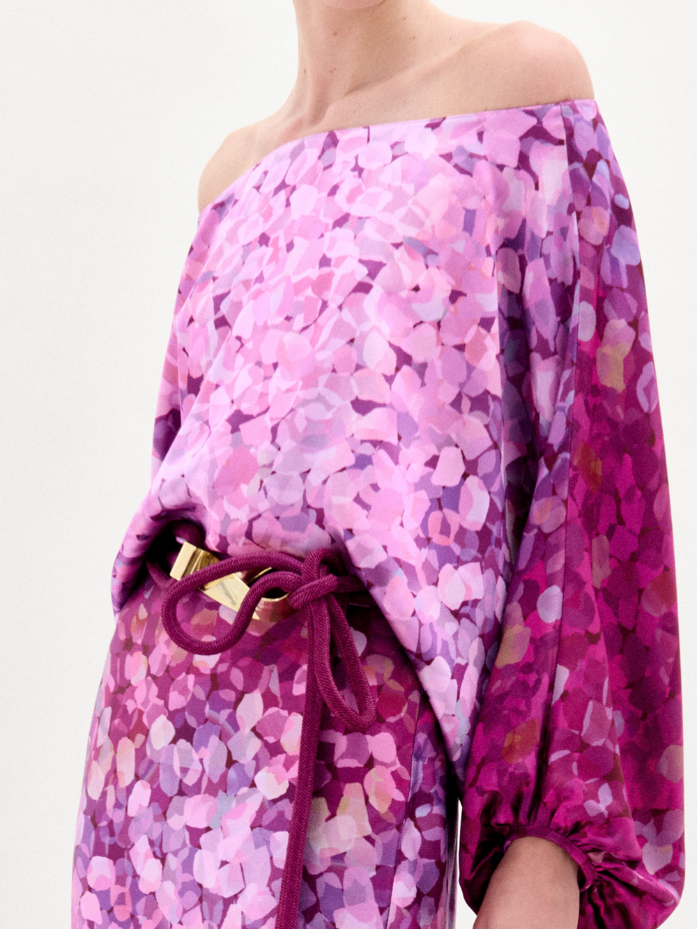 A person wearing the Lorella Dress, an off-the-shoulder maxi dress crafted from luxurious Italian fabric and adorned with a purple, pink, and magenta floral pattern, stands against a plain background.