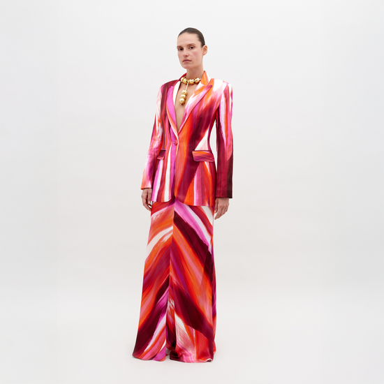 A person stands against a plain background, wearing a vibrant suit in red, pink, and orange streaks, paired with the Laurina Skirt Magenta Abstract Waves and bold gold necklace. This stunning ensemble will be available for pre-order soon and is set to ship by date 2024.