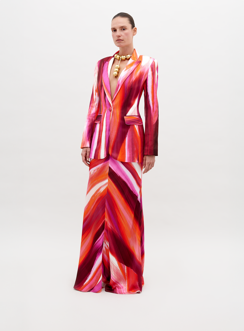 A person stands against a plain background, wearing a vibrant suit in red, pink, and orange streaks, paired with the Laurina Skirt Magenta Abstract Waves and bold gold necklace. This stunning ensemble will be available for pre-order soon and is set to ship by date 2024.