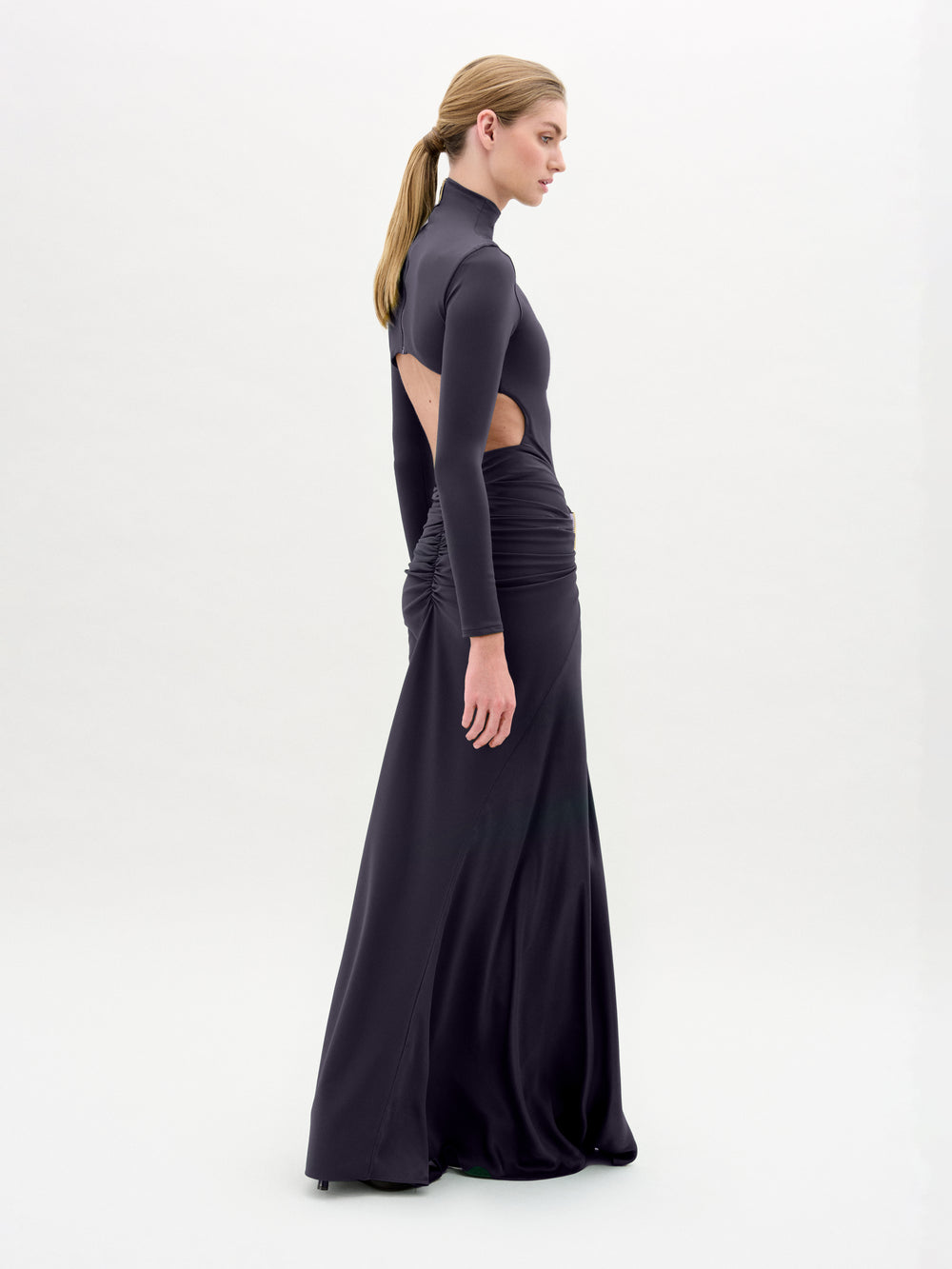 A woman wearing the Lena Dress—a long, fitted black dress with cut-out sides—stands in profile against a plain white background, her hair tied back in a low ponytail.