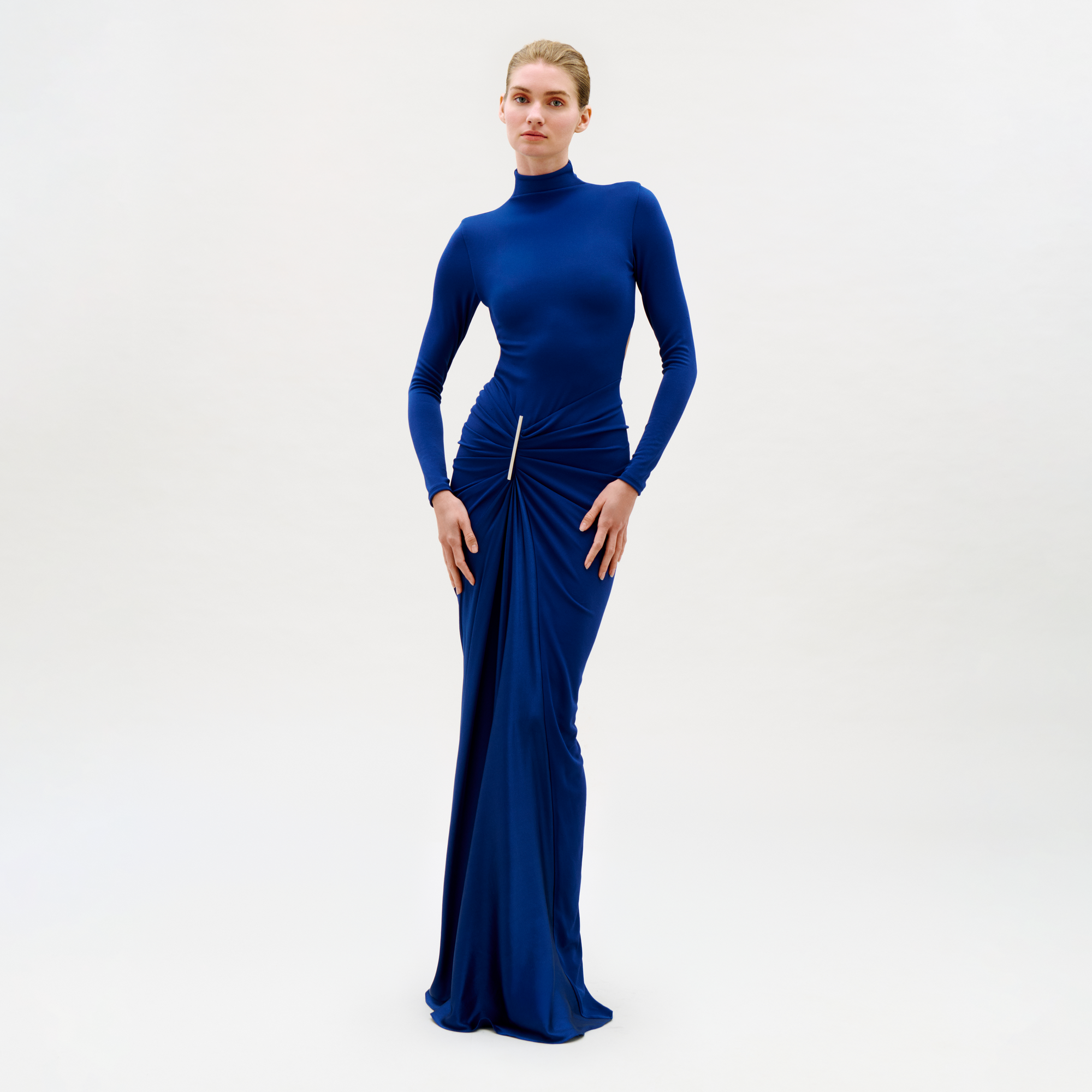 A person stands against a plain light background, wearing the Lena Dress—a long, fitted royal blue dress with long sleeves, high neckline, and gathered detail at the hip. Their elegant and confident pose highlights the dress’s sophisticated style.