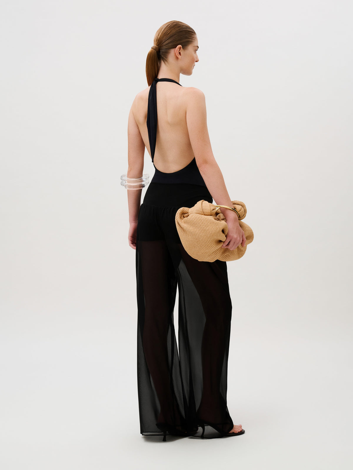A model viewed from behind wears a black halter-neck bodysuit and sheer chiffon Lene Pant, holding a large, textured beige clutch.