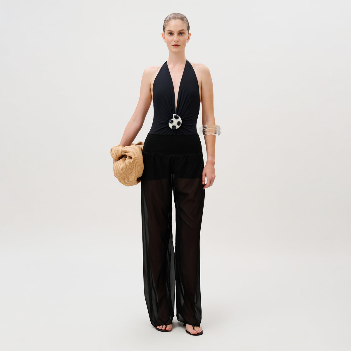 A model wears a black halter swimsuit with a large circular accent at the waist and sheer chiffon Lene Pant, carrying a tan woven clutch.