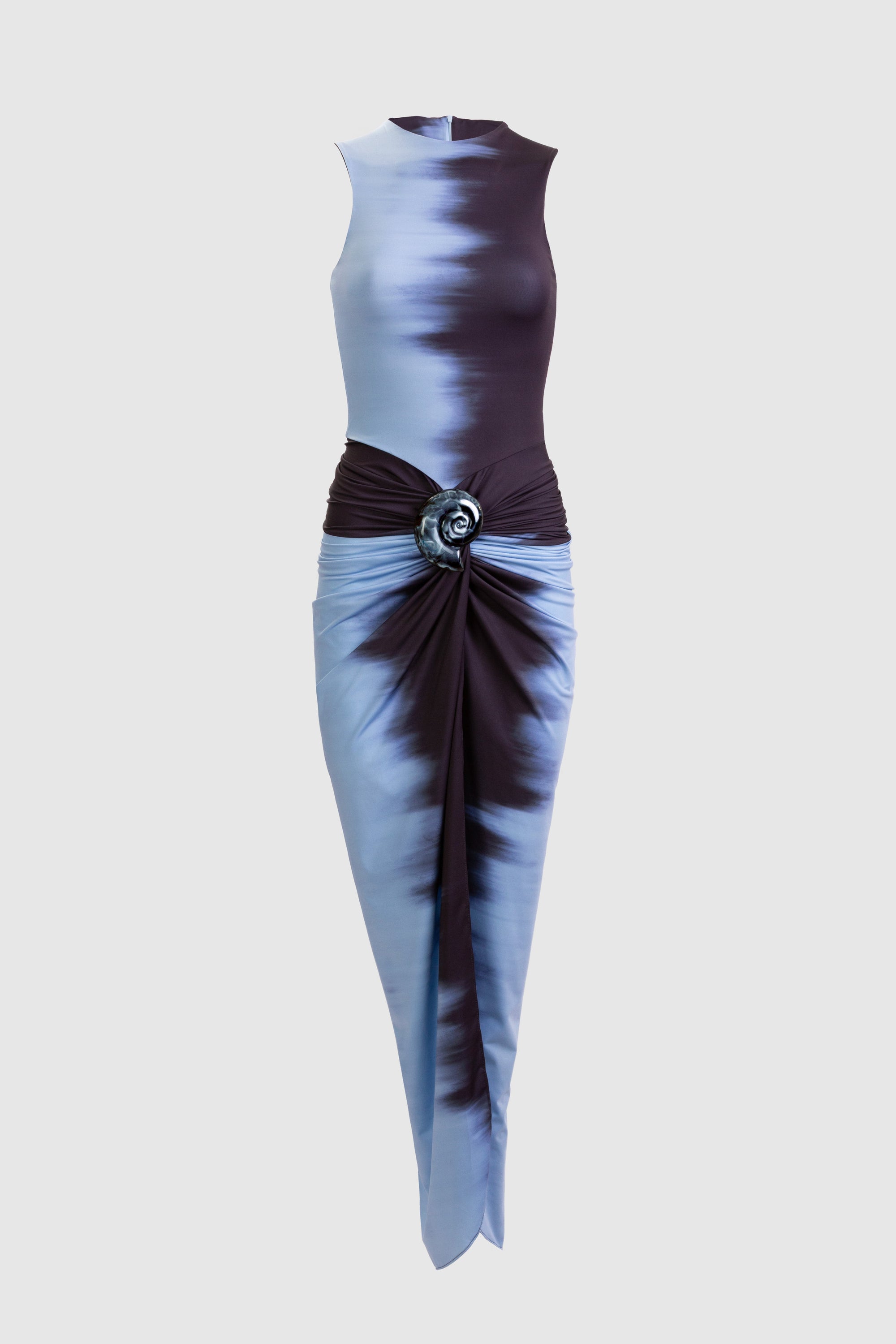 A woman wears the Leni Dress—a sleeveless blue satin dress with gathered detail and front slit—paired with a large black fringe handbag and black heeled sandals, standing against a plain background.