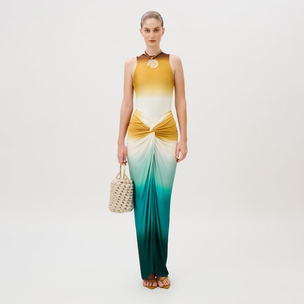 A person wears the Leni Dress, a slim-fit, floor-length piece in a yellow-to-teal ombre gradient with a twisted knot detail at the waist, carrying a cream woven handbag.