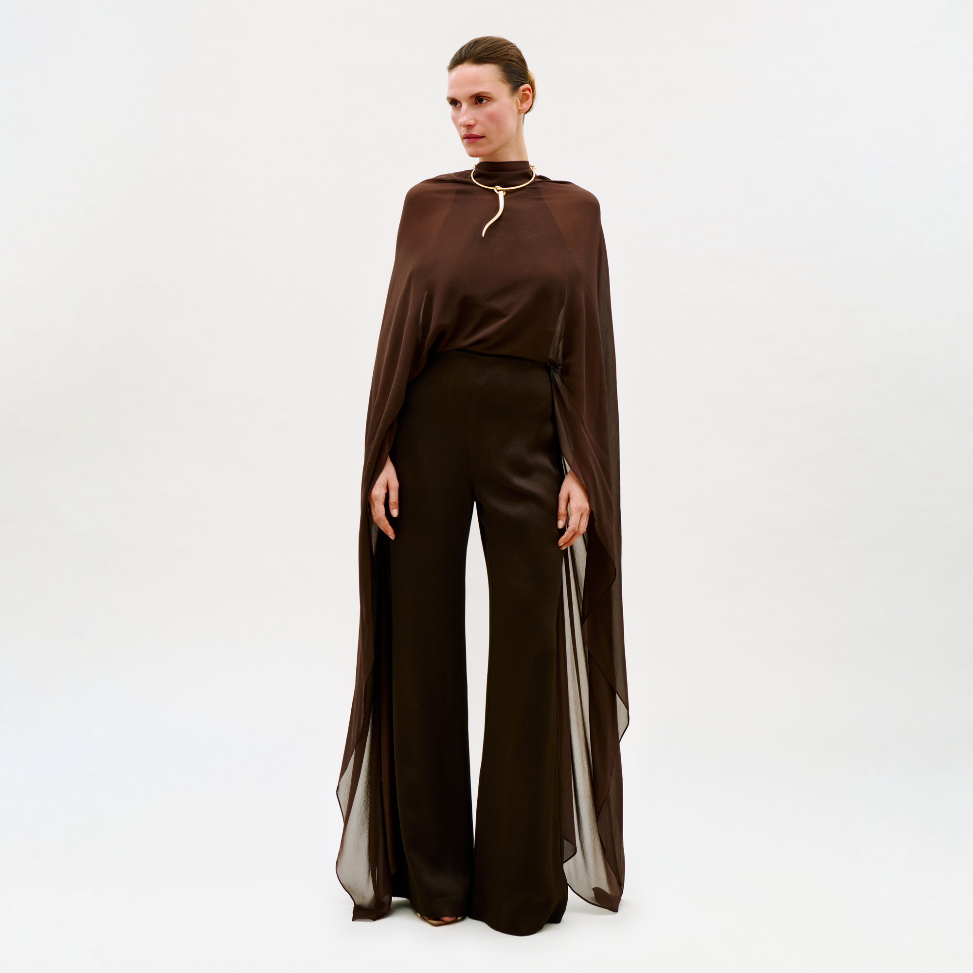 Against a plain white background, a woman wears the Ligia Jumpsuit—a dark brown, cape-like design with wide-leg trousers and sheer fabric draping elegantly from her shoulders to her ankles.