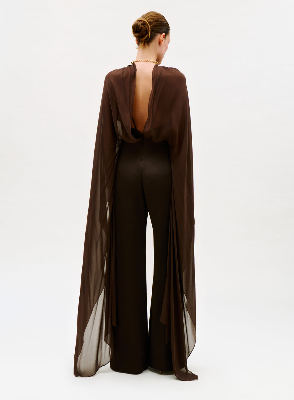 A person stands facing away, wearing the Ligia Jumpsuit—a sheer, flowing brown design with matching wide-leg pants and an open back—styled with a neat updo against a plain light background.