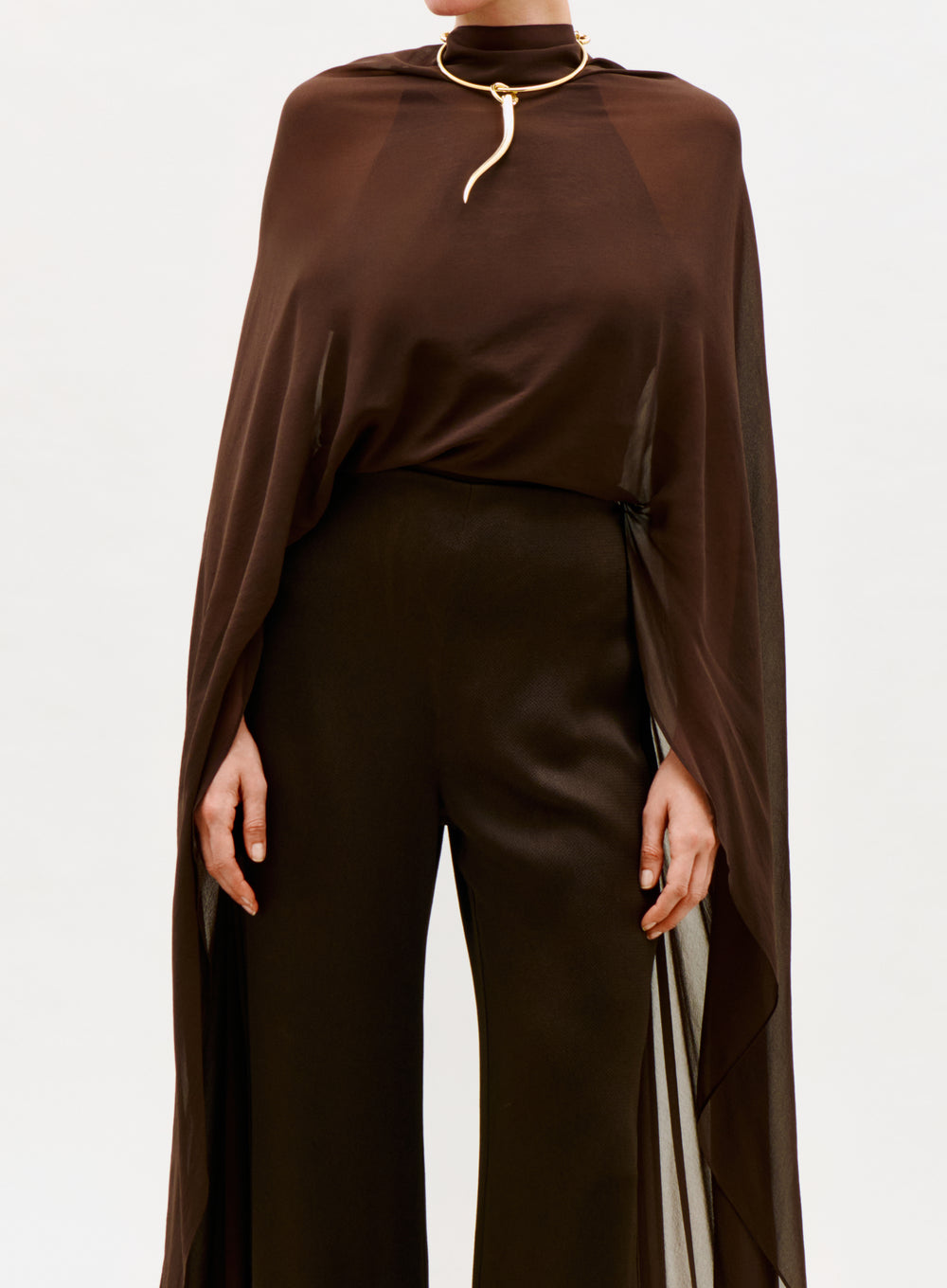 A person wears the Ligia Jumpsuit, a dark brown, semi-sheer piece with wide-leg pants, styled with a gold snake-shaped choker necklace. The background is plain white.