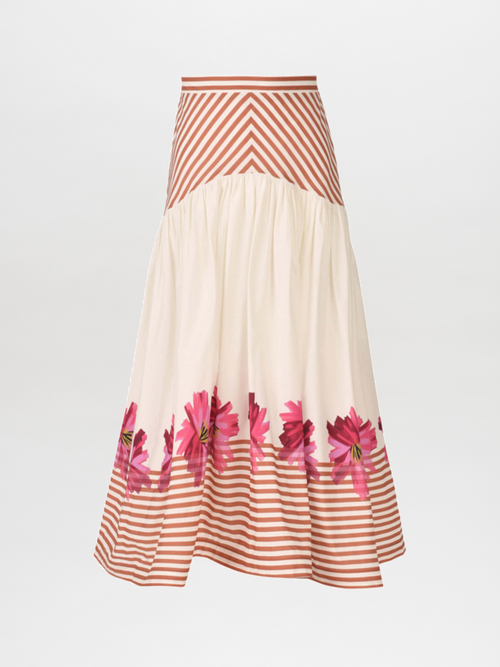 A woman poses against a plain backdrop wearing the Lilan Skirt in Magenta Floral Brushstroke Stripes, effortlessly complementing her sleeveless dress decorated with pink floral designs and orange stripes.