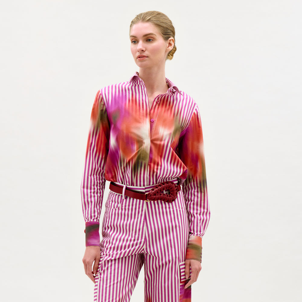 A woman stands against a white background wearing the Linette Blouse—a pink-and-white vertical striped long-sleeve top—paired with Italian fabric pants featuring red and green abstract details and a red floral belt accessory.