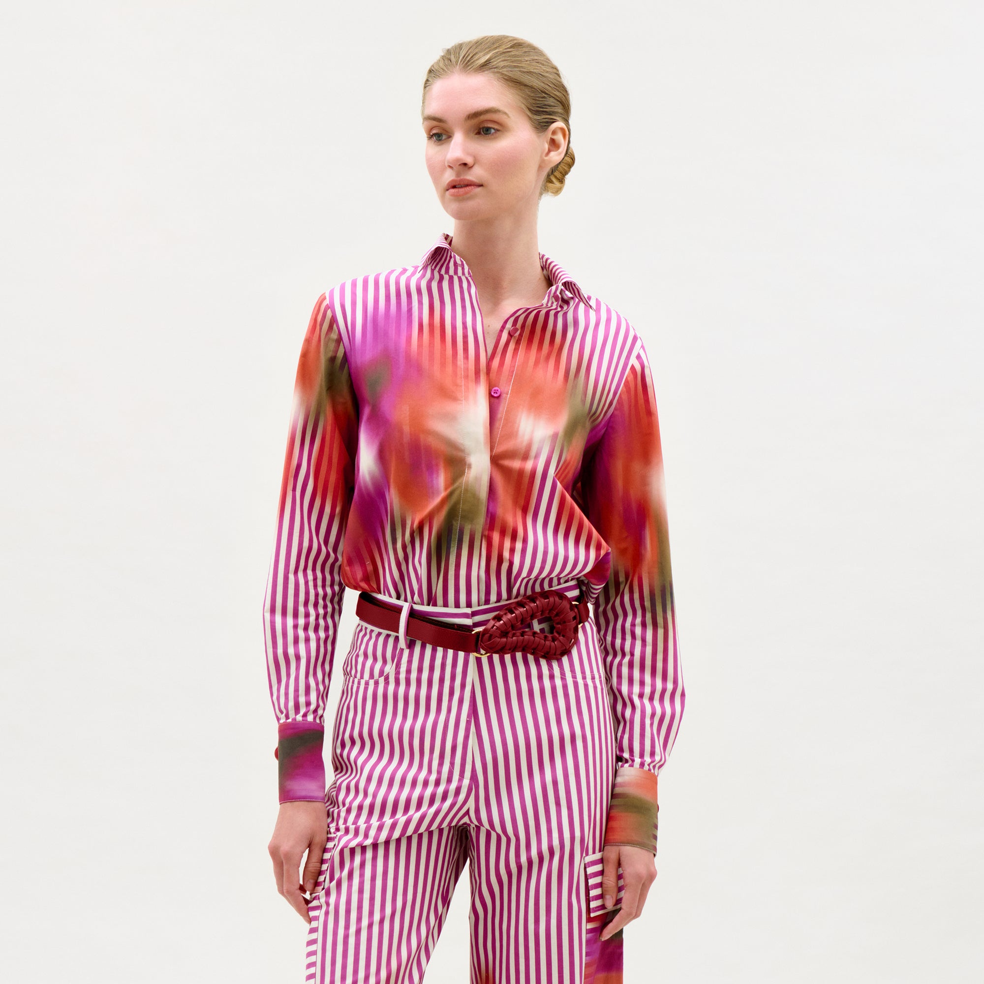 A woman stands against a white background wearing the Linette Blouse—a pink-and-white vertical striped long-sleeve top—paired with Italian fabric pants featuring red and green abstract details and a red floral belt accessory.