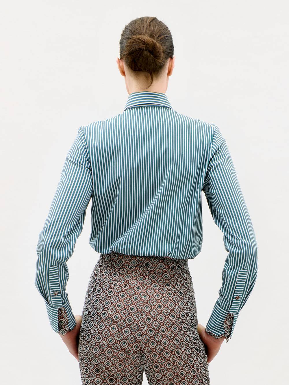 A person with brown hair in a bun faces away, wearing the Linnea Shirt—a blue and white vertical striped shirt—paired with patterned pants, against a plain white background.