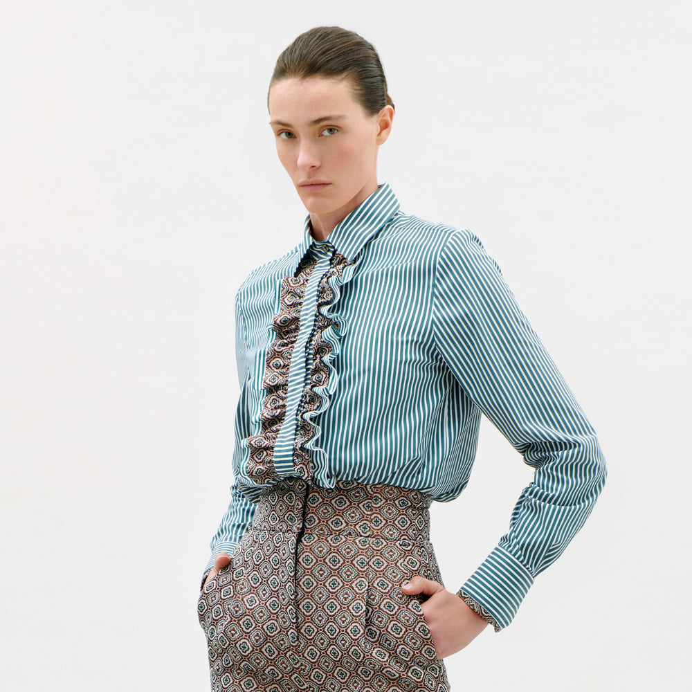 A person wearing the Linnea Shirt, a blue striped top with ruffles, and patterned high-waisted pants stands with hands in pockets against a plain white background.