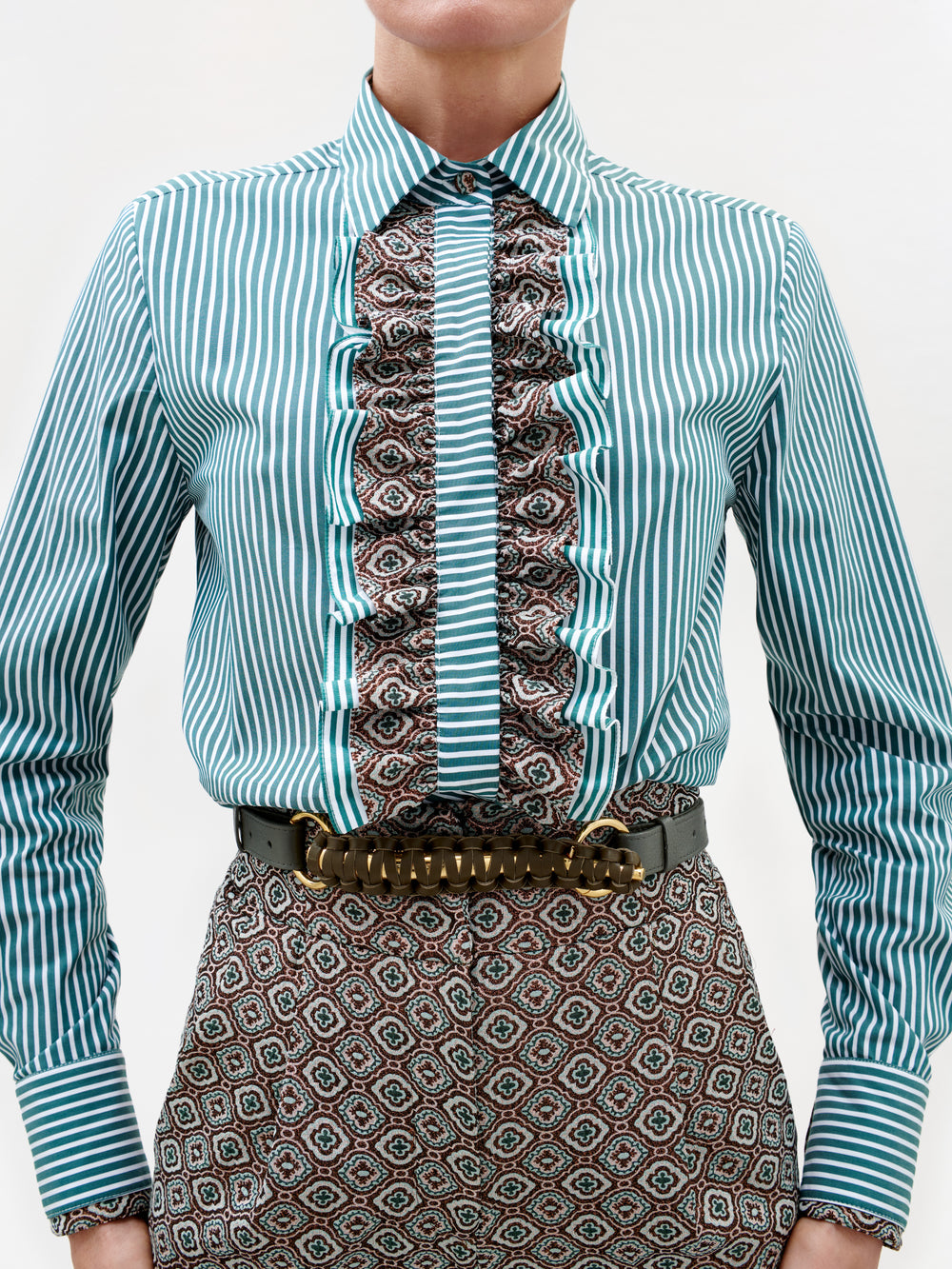 A person wearing the Linnea Shirt, featuring teal stripes, a ruffled center with mixed prints, paired with geometric patterned pants. A wide belt with a large gold buckle completes the outfit.