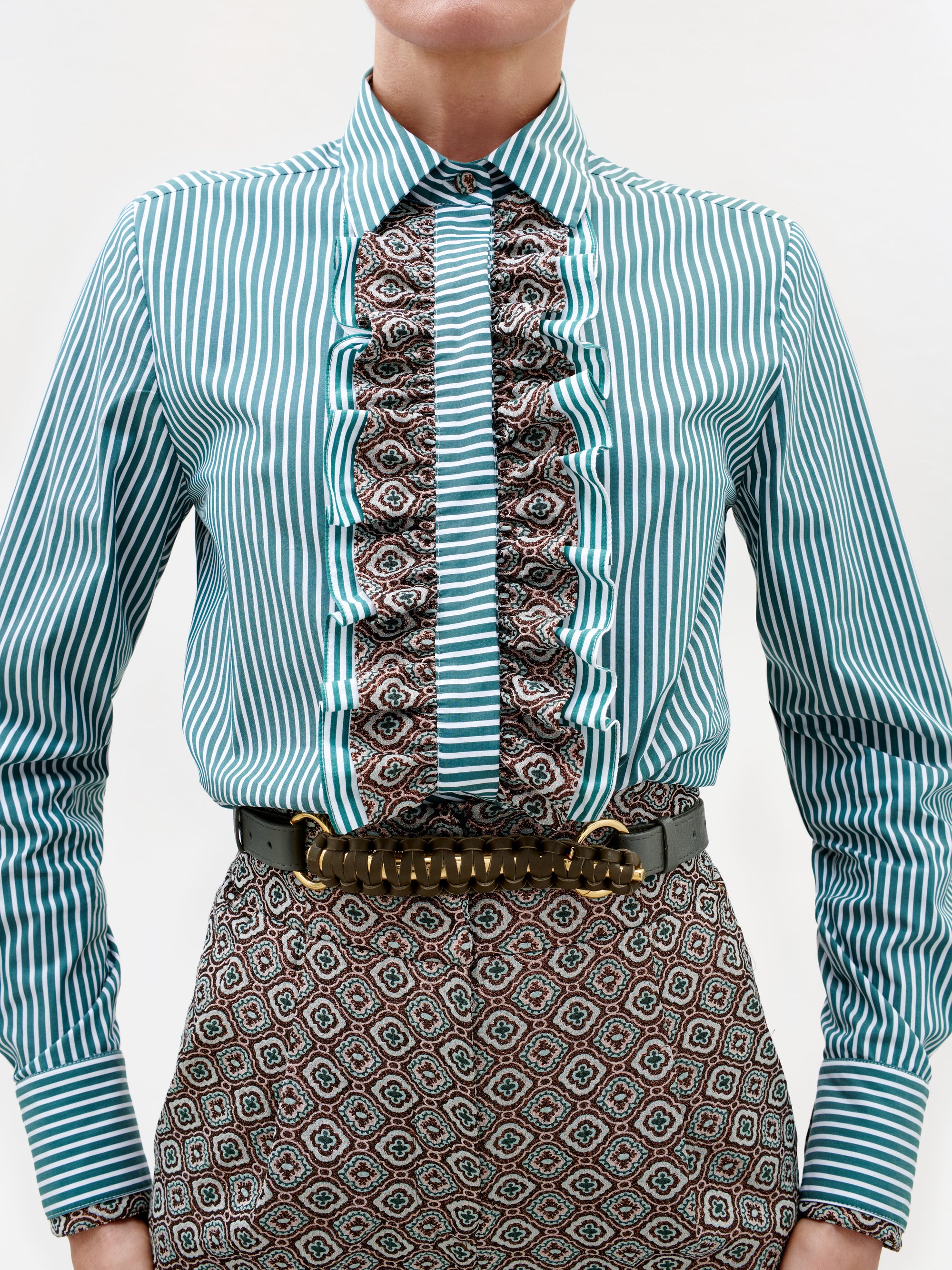 A person wearing the Linnea Shirt, featuring teal stripes, a ruffled center with mixed prints, paired with geometric patterned pants. A wide belt with a large gold buckle completes the outfit.