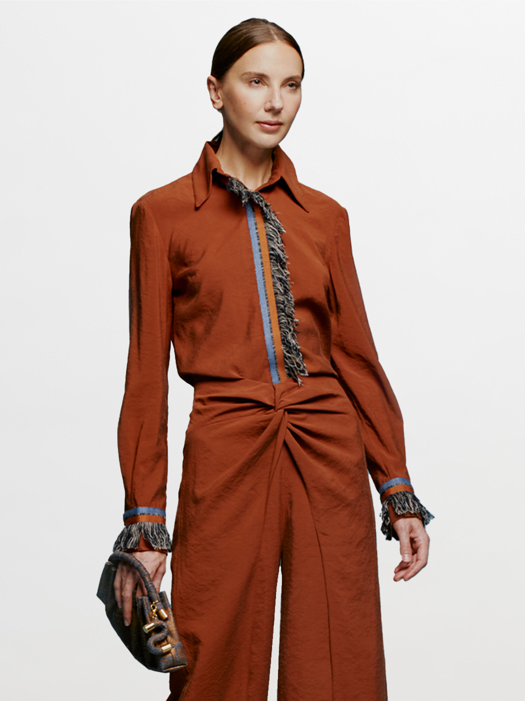 A person stands against a blue background wearing the Liv Blouse Cacao, a rust-colored outfit with fringe details on the collar, front, cuffs, and waistband, highlighting the latest trend expected in the September 2nd shipment. They're holding a small gray handbag.