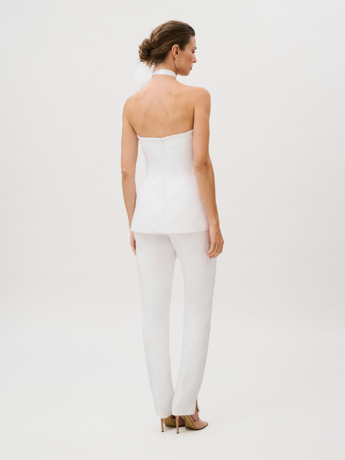 A woman stands facing away, styled in the Luca Pant—straight white pants paired with a white halter-top suit crafted from silk cotton brocade with textured fabric and finished with beige heels. Set against a plain light background, this elegant, ready-to-wear ensemble is made in Italy for sophisticated appeal.