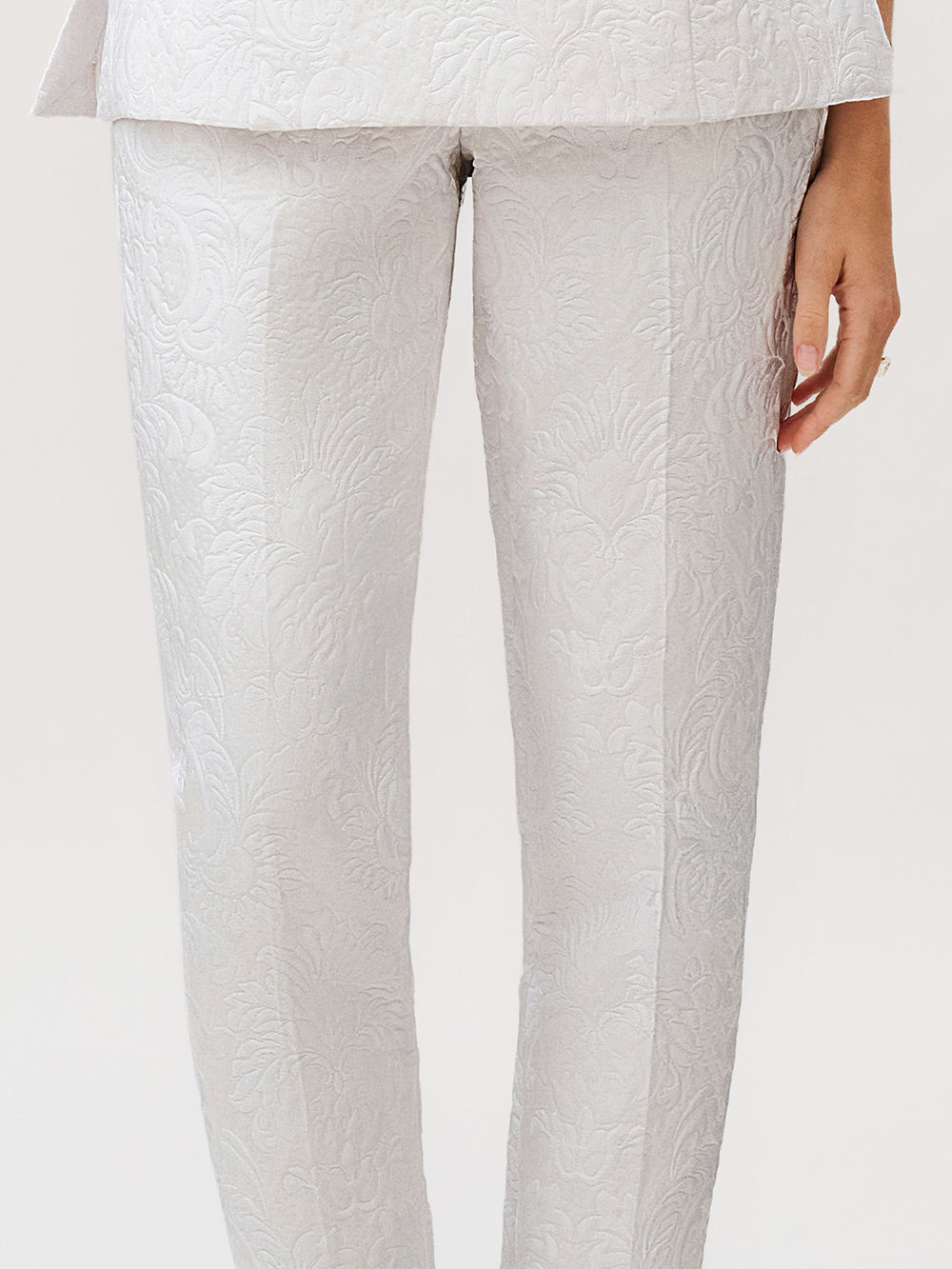 A person wearing the Luca Pant in light gray silk cotton brocade with a textured floral pattern, paired with a matching jacket—an elegant ready-to-wear ensemble made in Italy, with one hand visible by their side.