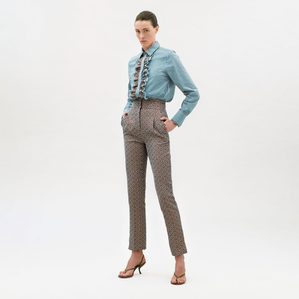A woman stands against a white background, wearing a blue striped blouse with ruffles and the Luca Pant—high-waisted, patterned pants crafted from exquisite Italian fabric. She has her hands in her pockets and is wearing heeled sandals.
