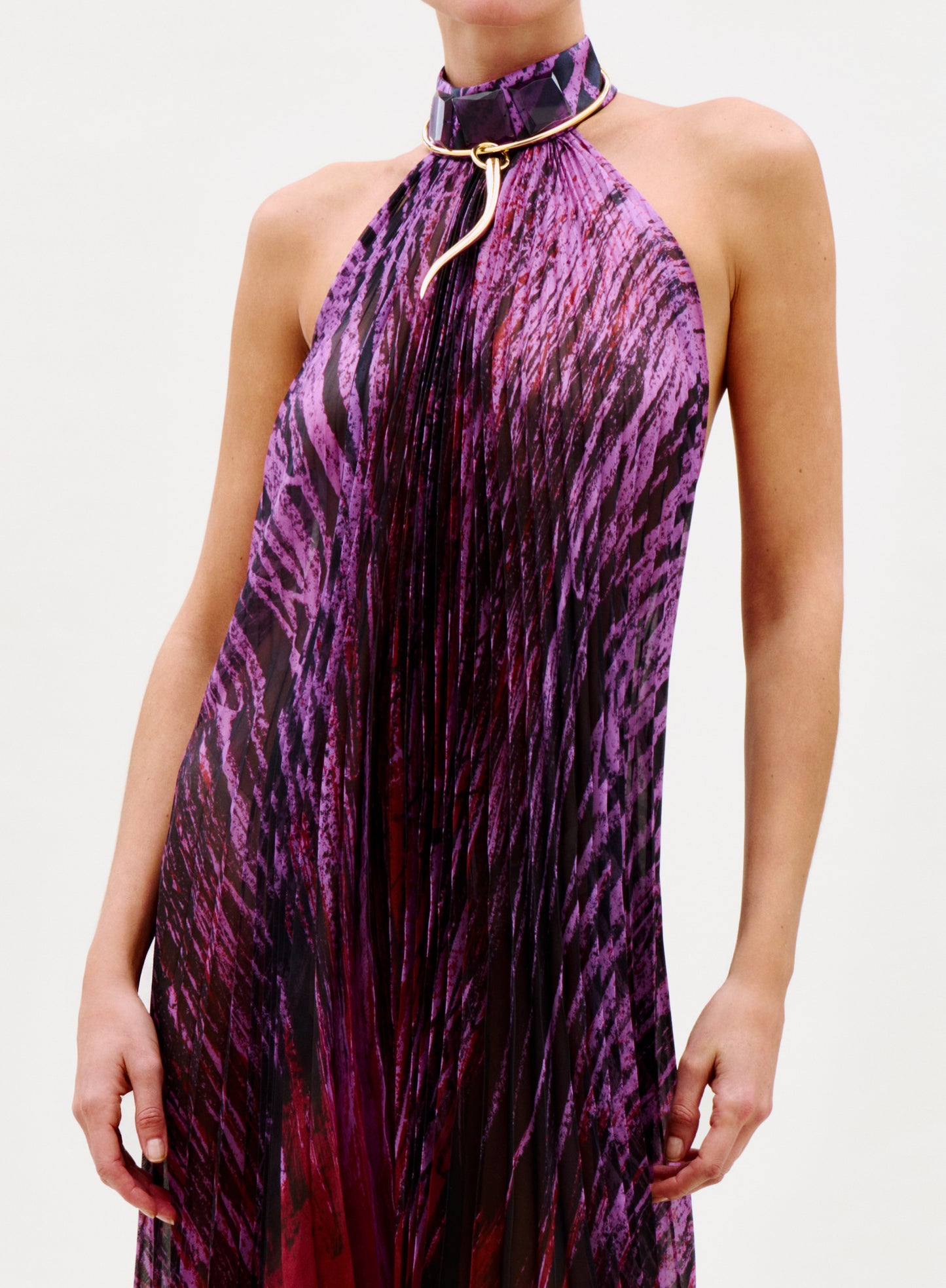 A person wearing the Lucia Dress—a sleeveless, pleated dress with a purple and black abstract pattern—and a matching choker with a gold metallic accent, set against a plain light-colored background.