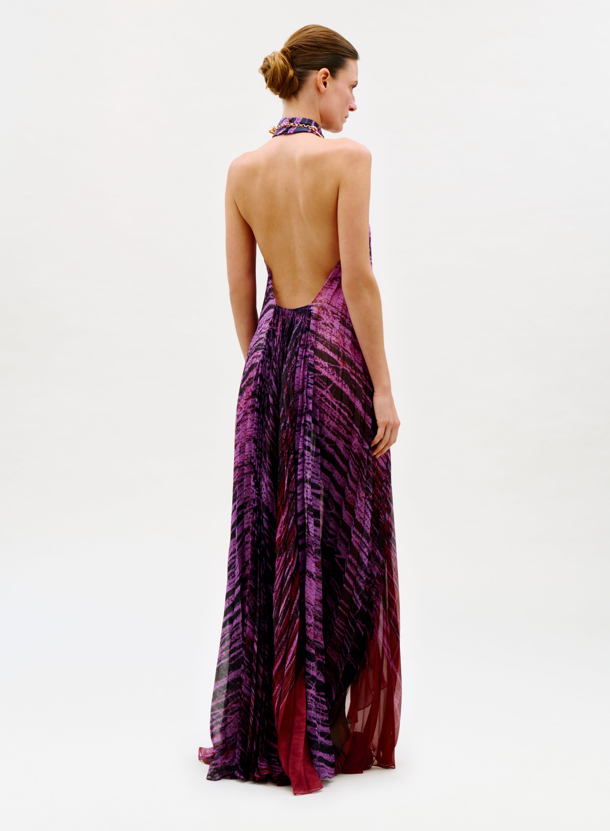 A woman stands facing away, wearing the Lucia Dress—a full-length, backless halter dress in purple and red abstract patterns. Her hair is styled in a neat bun against a plain white background.