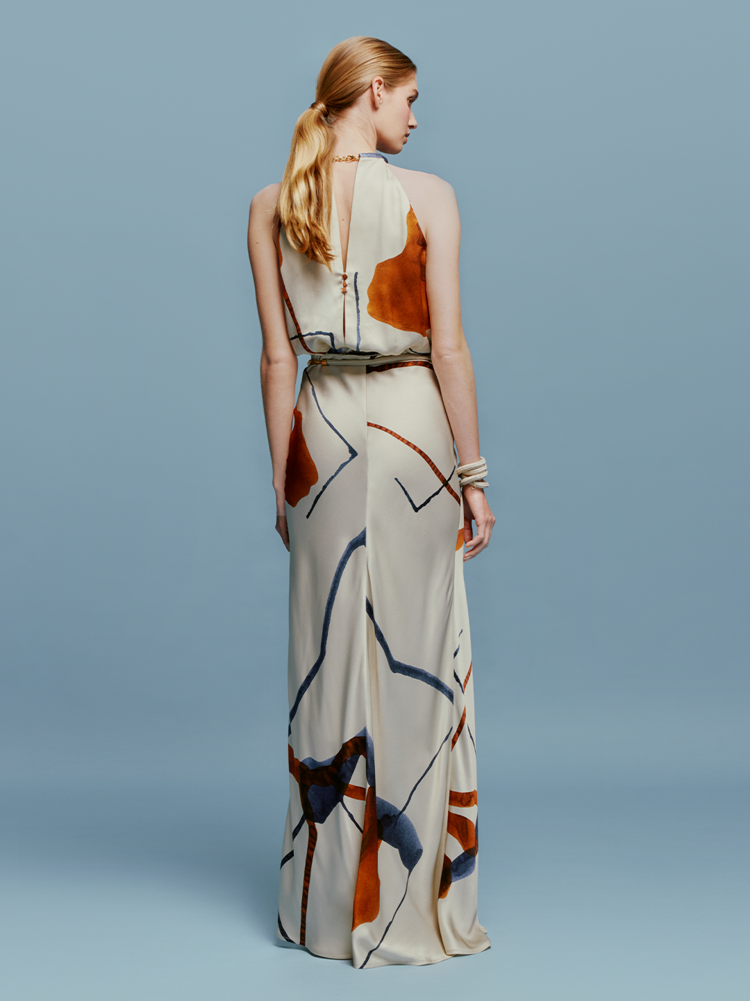 A woman stands against a blue background wearing the Manuela Skirt Cobalt Copper Abstract, a sleeveless, floor-length dress that features an intricate abstract pattern. She accessorizes with a bracelet on her wrist and gazes slightly to the side, effortlessly embodying elegance for August 19th, 2024.