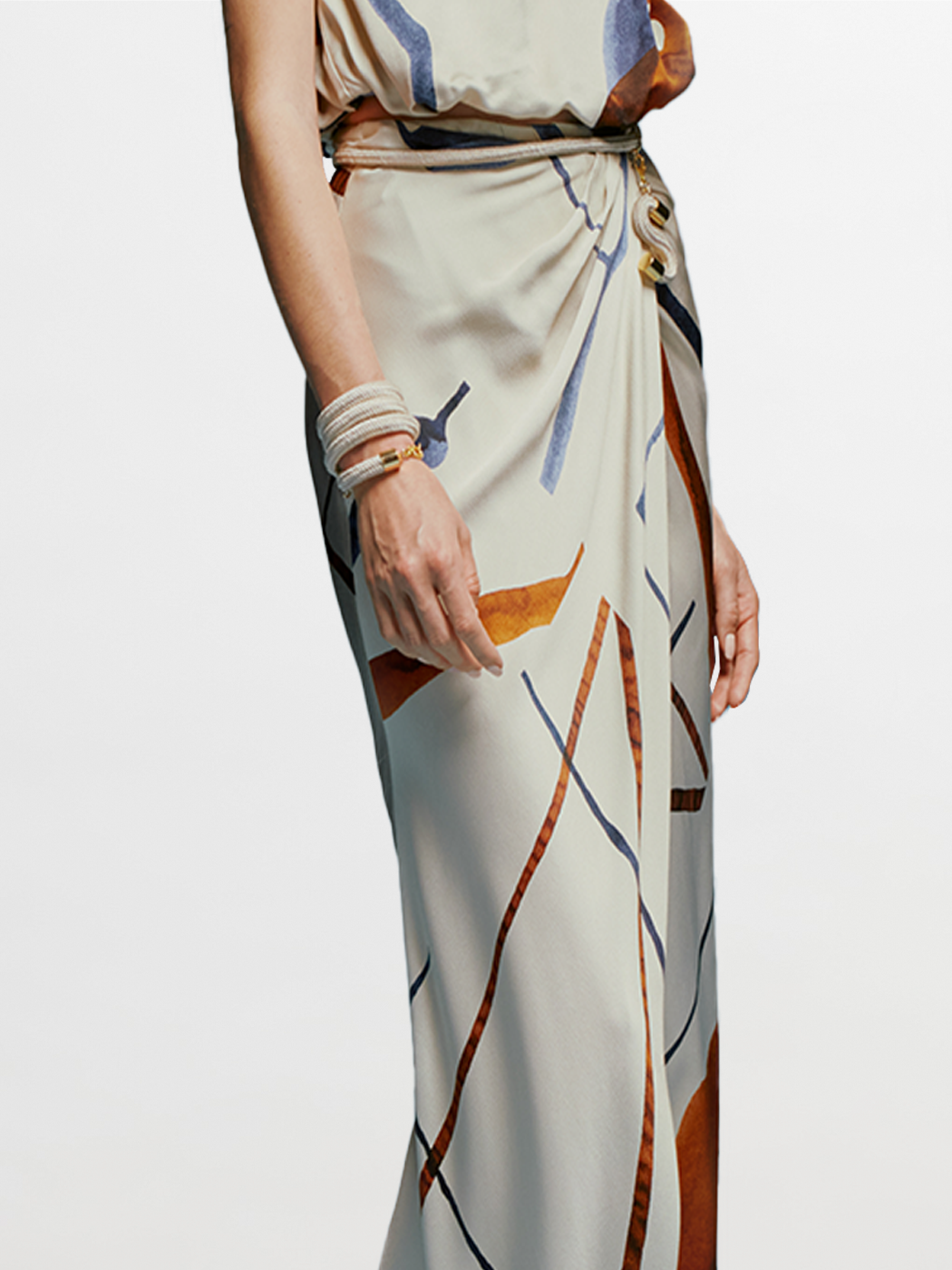 A woman stands against a blue background wearing the Manuela Skirt Cobalt Copper Abstract, a sleeveless, floor-length dress that features an intricate abstract pattern. She accessorizes with a bracelet on her wrist and gazes slightly to the side, effortlessly embodying elegance for August 19th, 2024.