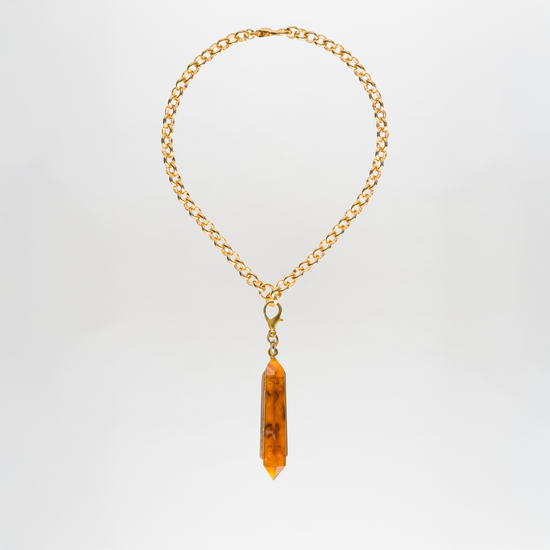 The Mariela Necklace is a gold chain adorned with a geometric crystal pendant, showcasing a striking elongated orange hue, beautifully displayed against a plain white background.